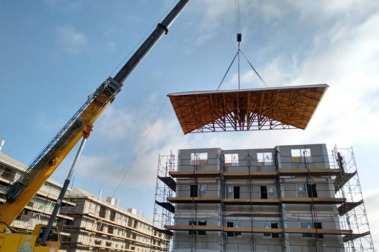 A imagem mostra um guindaste levantando uma estrutura de madeira em um canteiro de obras. Ao fundo, há um edifício em construção com andaimes ao redor. O céu está parcialmente nublado.
