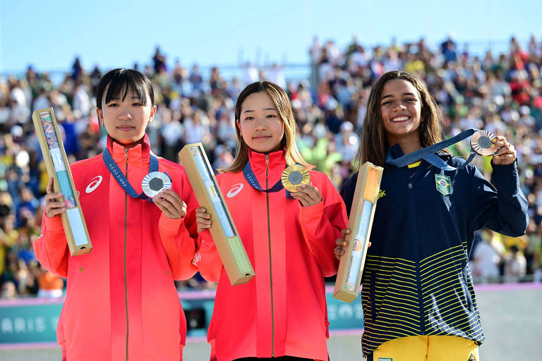 Três atletas estão em um pódio, segurando medalhas e prêmios. A atleta à esquerda usa uma jaqueta rosa e segura uma medalha prateada e um prêmio em forma de caixa. A atleta do meio, também vestindo rosa, segura um prêmio dourado e uma medalha prateada. A atleta à direita, vestindo uma jaqueta azul com detalhes amarelos, exibe um sorriso e segura uma medalha prateada e um prêmio. Ao fundo, uma multidão observa a cerimônia sob um céu claro.
