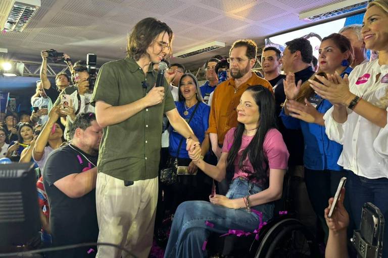 A imagem mostra um grupo de pessoas em um evento. Um homem de camiseta verde e calças claras está segurando um microfone e conversando com uma mulher em uma cadeira de rodas, que usa uma blusa rosa. Ao redor deles, várias pessoas estão aplaudindo e sorrindo, criando um ambiente de celebração. O fundo é decorado com bandeiras e cartazes.