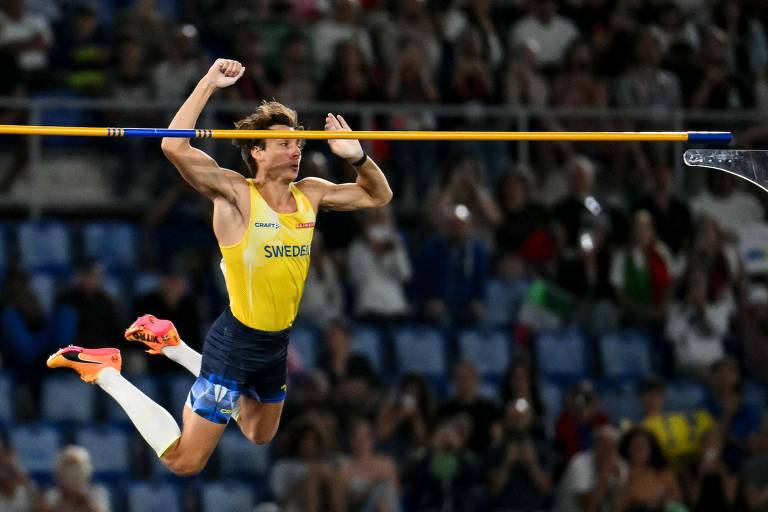 Um atleta está realizando um salto com vara, elevando-se acima de uma barra horizontal. Ele usa um uniforme amarelo e azul, com meias brancas. O fundo mostra uma multidão assistindo ao evento em um estádio.