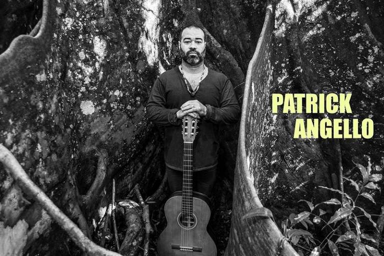 Em foto preto e branca, o violonista Patrick Angello posa para a câmera em meio à raízes de uma árvore