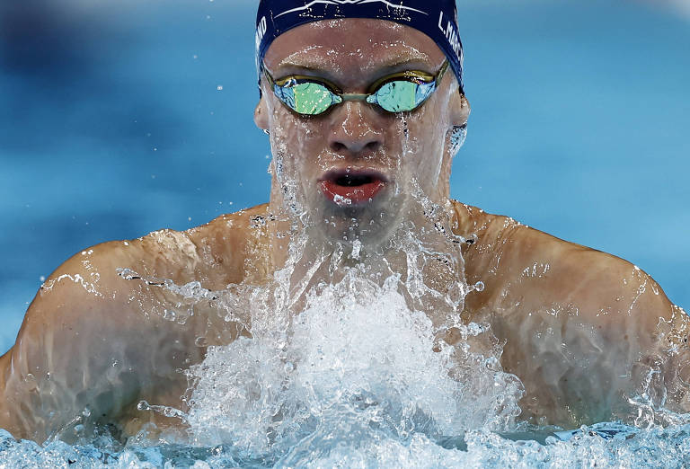 Um nadador está em movimento na água, com o rosto parcialmente submerso e respingos de água ao seu redor. Ele usa óculos de natação com lentes verdes e um touca de natação azul. O fundo é de uma piscina com água azul clara.