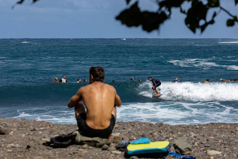A imagem mostra um homem sentado em uma pedra, de costas, observando surfistas no mar. O mar está agitado com ondas, e há várias pessoas surfando. O céu está claro e há algumas nuvens. O homem está sem camisa e usa calças curtas. Ao lado dele, há uma toalha de praia azul.