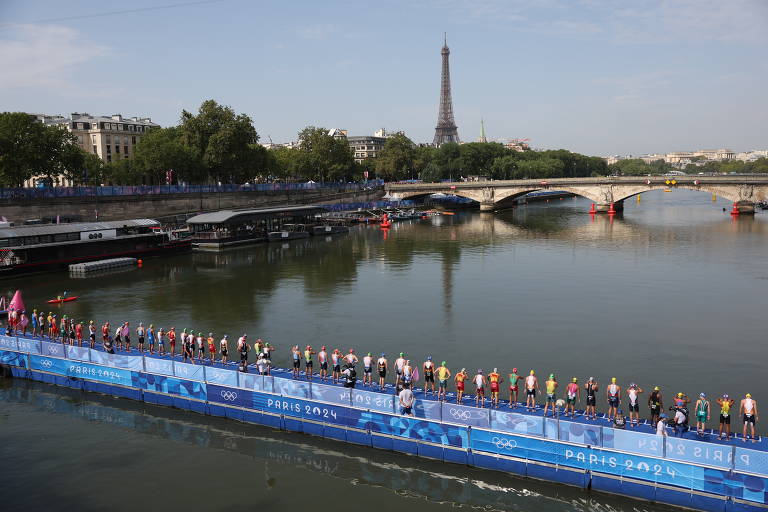 A imagem mostra uma vista do Rio Sena em Paris, com várias pessoas em uma plataforma flutuante azul. Ao fundo, é visível a Torre Eiffel e algumas árvores ao longo da margem do rio. O céu está claro e ensolarado, e há pontes que cruzam o rio
