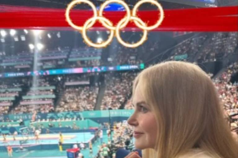 A imagem mostra uma competição de ginástica com várias atletas no palco. No fundo, há uma tela exibindo os resultados da final da equipe feminina, com a lista de países e suas pontuações. Uma mulher de cabelo loiro e liso, vestindo uma blusa clara, está sentada na plateia, observando a competição. O ambiente está cheio de espectadores, e há bandeiras visíveis, incluindo uma do Brasil.