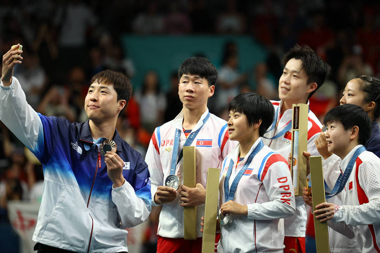 Um grupo de seis atletas está posando para uma selfie em um evento esportivo. Eles estão sorrindo e segurando medalhas e prêmios. O ambiente ao fundo é de uma competição, com uma multidão visível. Os atletas estão vestidos com uniformes esportivos, predominantemente brancos com detalhes em vermelho e azul.