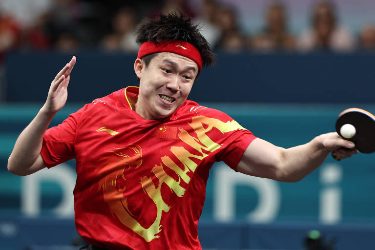 Um jogador de tênis de mesa está em movimento, usando uma camiseta vermelha com a palavra 'CHINA' em destaque. Ele tem um headband vermelho e está segurando uma raquete de tênis de mesa, parecendo concentrado e animado durante a partida. O fundo mostra uma multidão, sugerindo um evento esportivo.
