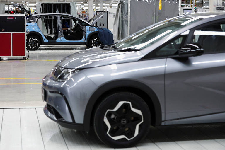 A imagem mostra um carro cinza em primeiro plano, com um design moderno e rodas de formato distinto. Ao fundo, há um carro azul parcialmente montado, visível em uma linha de produção. O ambiente é uma fábrica, com equipamentos e ferramentas visíveis ao redor.