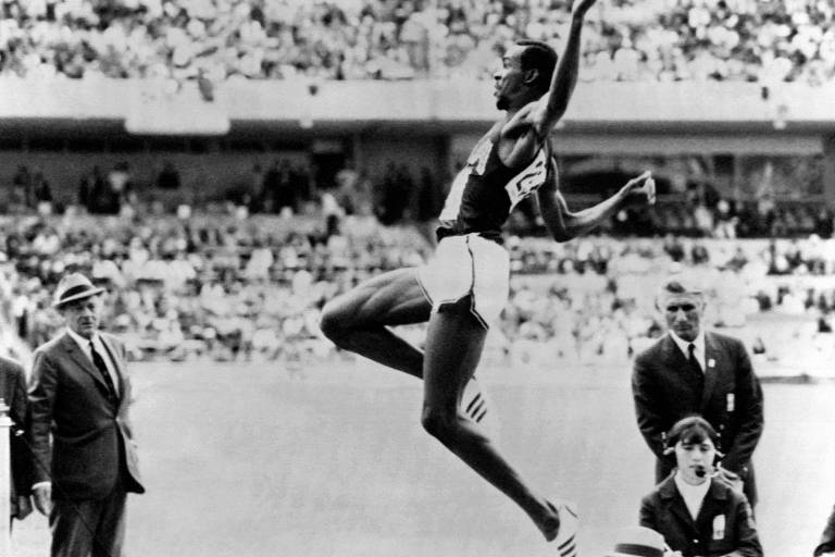 A imagem em preto e branco mostra um atleta realizando um salto em distância em um estádio lotado. O atleta está no ar, com os braços levantados e as pernas estendidas. Ao fundo, há uma multidão de espectadores. À esquerda, dois homens em trajes formais observam, enquanto uma mulher com fones de ouvido está sentada à direita, em uma mesa.
