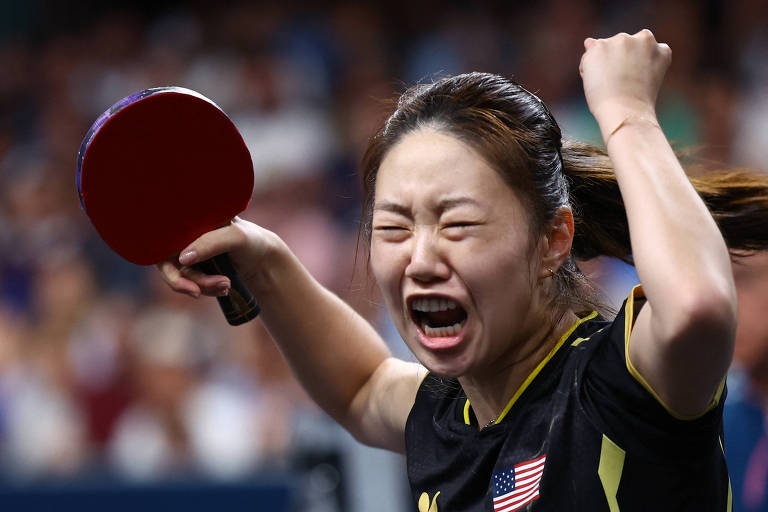 Uma jogadora de tênis de mesa está comemorando com um gesto de vitória. Ela segura uma raquete vermelha e parece estar gritando de alegria. A atleta está vestindo uma camisa preta com detalhes em amarelo e um emblema dos Estados Unidos. O fundo mostra uma multidão, sugerindo um ambiente de competição.
