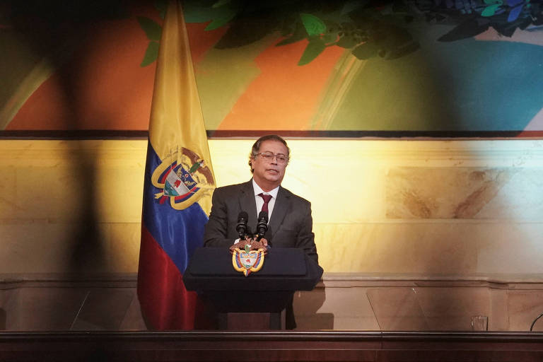 Um homem em um terno escuro está em pé atrás de um púlpito, falando ao microfone. Ao fundo, há uma bandeira da Colômbia, com as cores amarela, azul e vermelha. O ambiente é bem iluminado, com uma decoração que sugere um evento formal.
