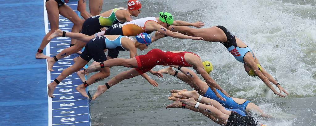A imagem mostra vários atletas em trajes de natação saltando simultaneamente para a água. Eles estão posicionados em uma plataforma de largada, com os corpos inclinados para frente e os braços estendidos. Os atletas usam toucas de diferentes cores, como vermelho, verde e amarelo, e a água parece agitada, sugerindo um ambiente de competição.