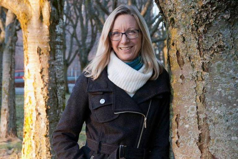A imagem mostra Elisabeth Oxfeldt, uma mulher sorridente, com óculos e cabelo liso, vestindo um casaco preto e um cachecol claro. Ela está encostada em uma árvore em um ambiente ao ar livre, com árvores ao fundo e uma iluminação suave, sugerindo um momento tranquilo.