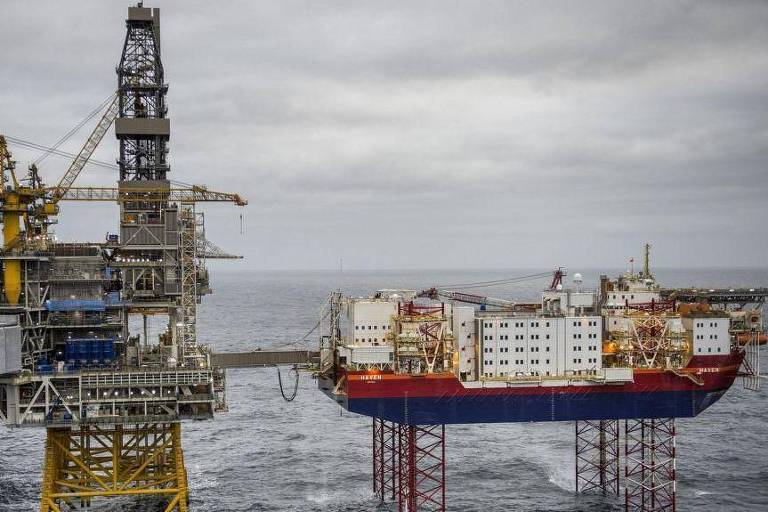 A imagem mostra duas plataformas de perfuração no mar. À esquerda, uma plataforma alta com uma torre de perfuração e estruturas de suporte. À direita, uma plataforma flutuante com um casco de navio, ancorada sobre pilares. O céu está nublado e o mar parece agitado.