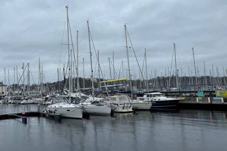 A imagem mostra uma marina com vários barcos ancorados. O céu está nublado, e a água reflete a luz cinza. Ao fundo, há um edifício de vários andares, e os mastros dos barcos se destacam contra o céu. A cena transmite uma atmosfera tranquila e serena.