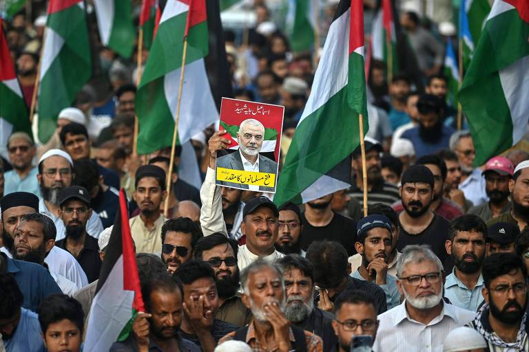 A imagem mostra uma grande multidão de pessoas segurando bandeiras palestinas. No centro, um homem levanta um cartaz com uma foto de um líder e texto em árabe. A multidão é composta por homens e mulheres de diferentes idades, muitos com expressões sérias, demonstrando apoio à causa palestina.