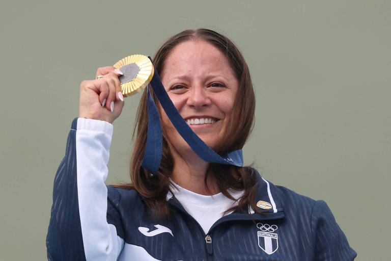 Uma atleta sorri enquanto segura uma medalha de ouro acima da cabeça. Ela está vestindo uma jaqueta esportiva azul e branca. O fundo é de uma cor sólida, possivelmente verde.
