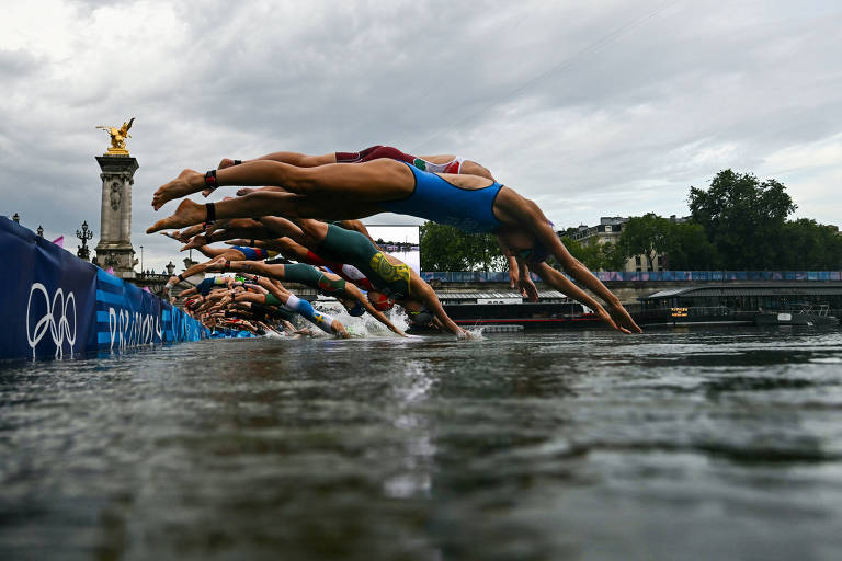 A imagem mostra vários atletas mergulhando na água de uma competição, com os corpos inclinados para frente e as mãos estendidas. Ao fundo, é possível ver uma estrutura arquitetônica e um céu nublado.
