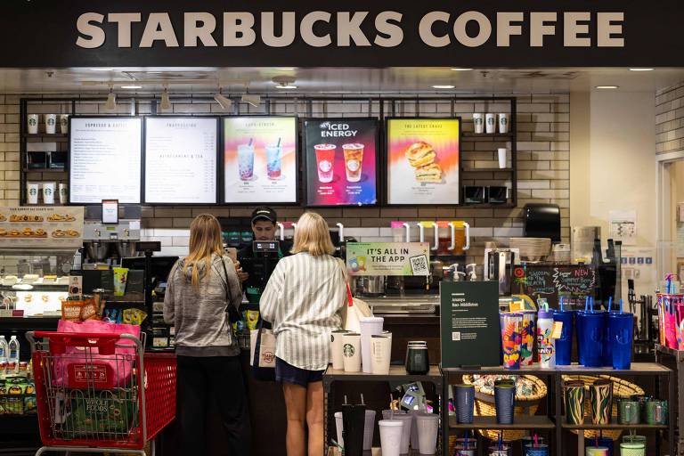 A imagem mostra duas pessoas em pé em frente ao balcão de um café Starbucks. Acima do balcão, há uma placa com o nome 'STARBUCKS COFFEE'. No fundo, há telas com opções de bebidas e produtos expostos em prateleiras. O ambiente é iluminado e organizado, com uma variedade de itens de café e acessórios visíveis.