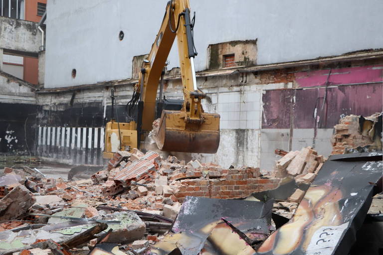 A imagem mostra uma máquina de demolição, possivelmente uma escavadeira, em ação, derrubando uma estrutura de tijolos. Há destroços de tijolos e materiais de construção espalhados pelo chão, com uma parede de fundo parcialmente visível, onde se pode notar algumas janelas e uma pintura em tons de rosa.