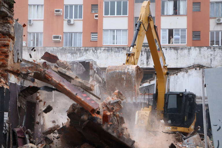 A imagem mostra uma máquina de demolição, com um braço mecânico, derrubando uma estrutura de um edifício. Há destroços e poeira sendo levantados, enquanto o fundo apresenta janelas de um prédio residencial. A cena retrata um processo de demolição em andamento.