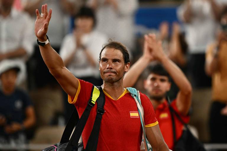 Um jogador de tênis, vestindo uma camiseta vermelha com detalhes amarelos, acena para a multidão enquanto é aplaudido. Ele parece emocionado e está em um ambiente de competição, com espectadores ao fundo. O jogador carrega uma mochila nas costas.
