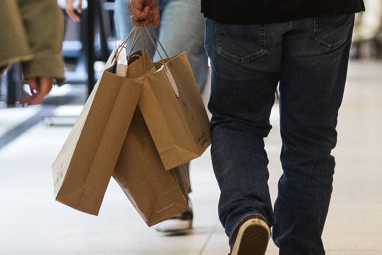 A imagem mostra um ambiente de shopping com várias pessoas. À esquerda, uma mulher jovem está usando uma blusa verde e shorts, olhando para o celular. No centro, um homem de costas, vestindo uma camisa preta e jeans, segura sacolas de compras. Ao lado dele, uma mulher com cabelo solto e vestido branco está segurando a mão dele. Ao fundo, outras pessoas estão caminhando e há mesas de um restaurante. Todos estão usando máscaras.