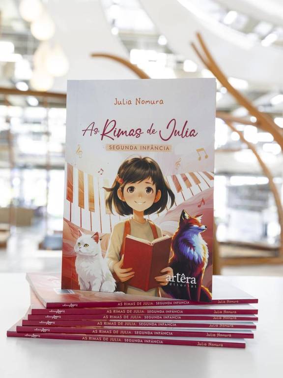 A imagem mostra a capa do livro 'As Rimas de Julia', escrito por Julia Nomura. A capa apresenta uma ilustração de uma menina sorridente segurando um livro, com um gato branco e uma raposa ao seu lado. Ao fundo, há um piano e notas musicais flutuando. O livro está em destaque sobre uma pilha de cópias do mesmo título, com o texto 'AS RIMAS DE JULIA: SEGUNDA INFÂNCIA' visível na parte inferior.