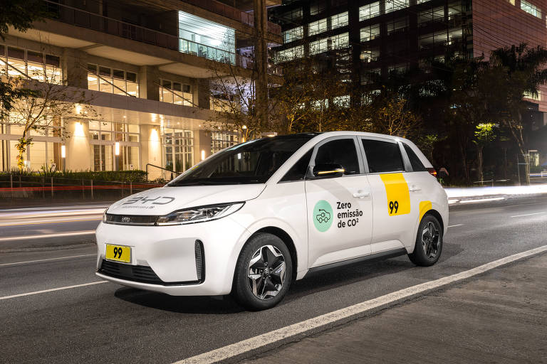 Carro branco com logo "BYD" no capô andando em rua durante a noite. Atrás dele, prédios iluminados. 