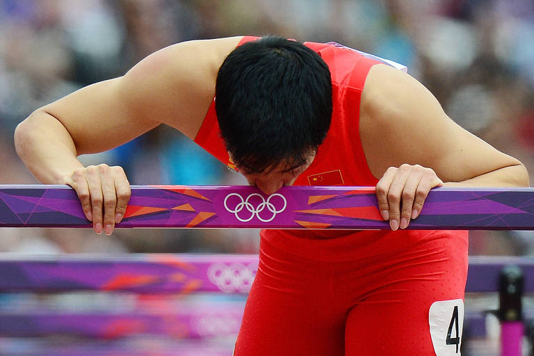 Um atleta vestido com um uniforme vermelho está se inclinando sobre uma barra de obstáculos, aparentemente exausto ou concentrado. O fundo mostra uma multidão, sugerindo que a imagem foi capturada durante uma competição olímpica.