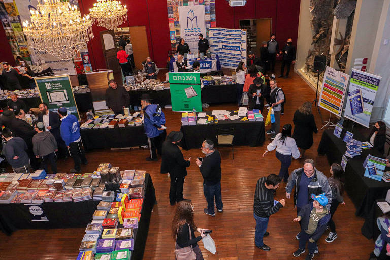 A imagem mostra uma feira de livros em um ambiente interno, com várias mesas dispostas em um espaço amplo. Há pessoas circulando entre as mesas, algumas conversando e outras examinando os livros expostos. As mesas estão cobertas com livros de diferentes tamanhos e cores, e há banners e cartazes visíveis ao fundo, promovendo diferentes editoras e produtos. Um grande lustre está pendurado no teto, iluminando o espaço.