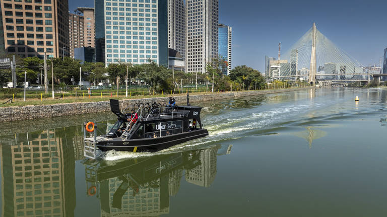 A imagem mostra um barco preto navegando no Rio Pinheiros, em São Paulo. Ao fundo, há arranha-céus e a famosa ponte Estaiada. O céu está claro e o reflexo do barco e dos edifícios na água é visível.