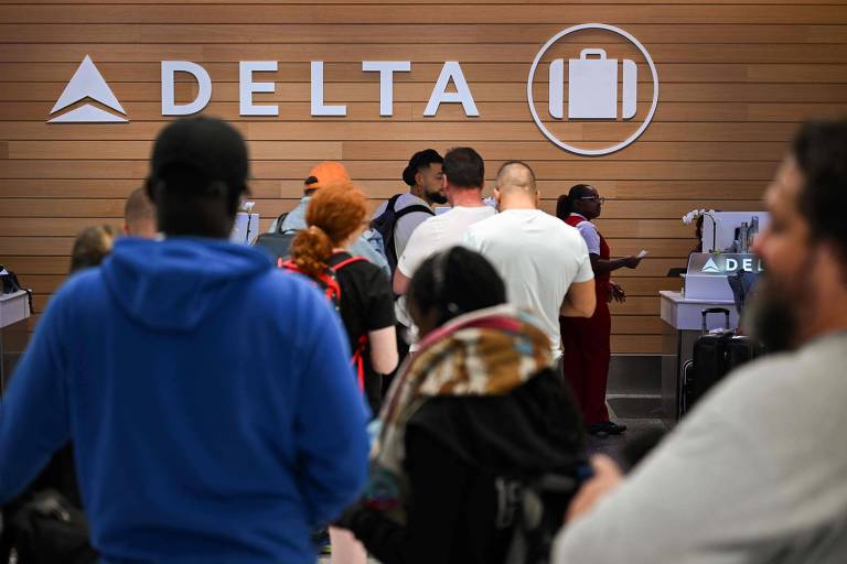 A imagem mostra uma fila de pessoas em frente ao balcão da Delta Airlines em um aeroporto. O logotipo da Delta está visível na parede ao fundo, junto a um ícone de mala. Algumas pessoas estão de costas, enquanto outras estão sendo atendidas por funcionários da companhia aérea. O ambiente é moderno, com um design de madeira clara.
