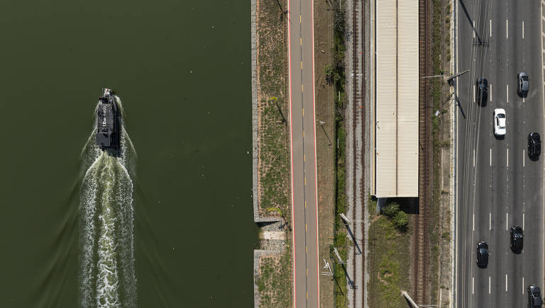 A imagem mostra uma vista aérea de um canal com um barco navegando em direção à parte inferior da imagem. À direita, há uma estrada com vários carros em movimento, e à esquerda, uma calçada e uma linha de trem. O canal é de cor verde escura, e o barco deixa um rastro na água.