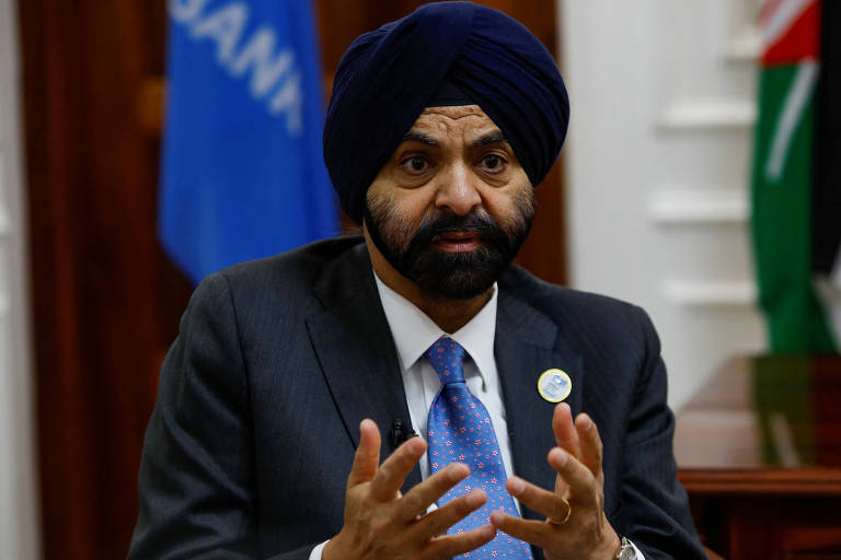 Um homem com um turbante azul e barba está sentado em uma mesa, gesticulando com as mãos enquanto fala. Ele usa um terno escuro e uma gravata azul. Ao fundo, há bandeiras visíveis, incluindo uma bandeira azul e outra com as cores branco e verde.
