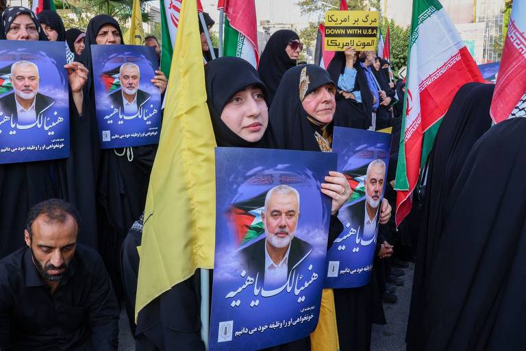 A imagem mostra um grupo de pessoas em um protesto, a maioria vestindo roupas pretas e as mulheres cobrindo a cabeça com o véu islâmico. Elas seguram cartazes com a imagem de Haniyeh e bandeiras do Irã.