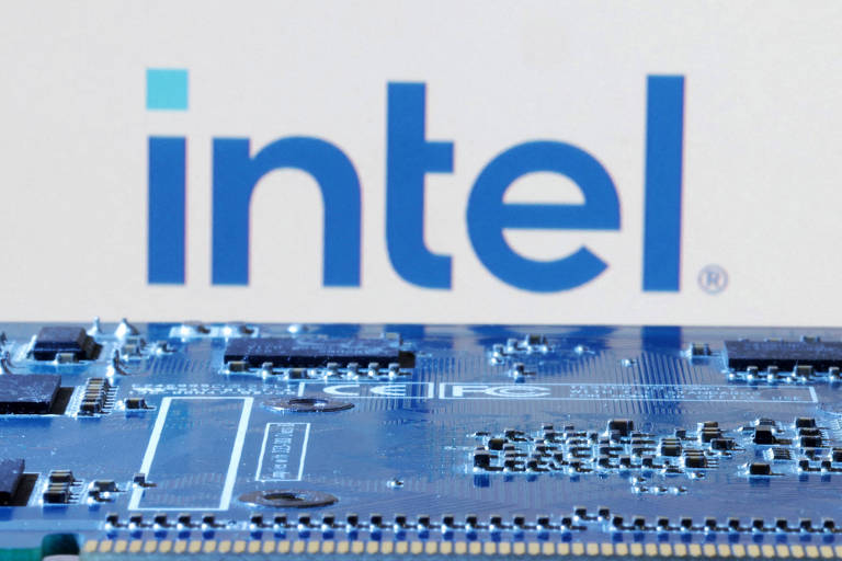A imagem mostra uma placa de circuito eletrônico em primeiro plano, com vários componentes eletrônicos visíveis, como chips e conectores. Ao fundo, está o logotipo da Intel em letras grandes e azuis, sobre um fundo claro.
