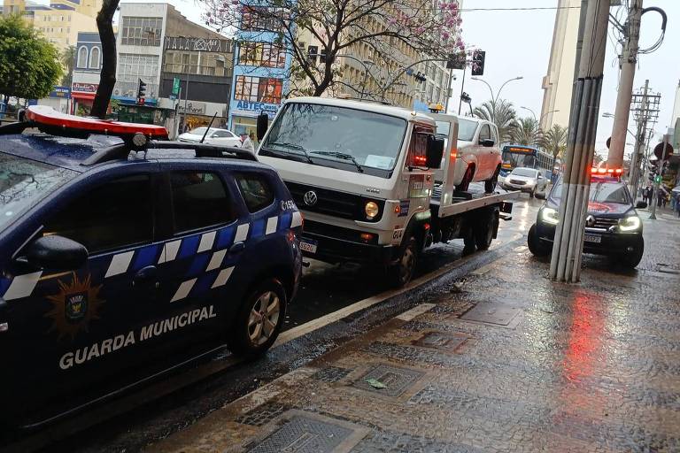 A imagem mostra uma cena urbana chuvosa com um caminhão guincho branco carregando um veículo. À esquerda, há um carro da Guarda Municipal com a inscrição 'GUARDA MUNICIPAL' e listras azuis e brancas