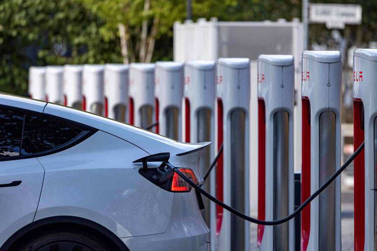 A imagem mostra a traseira de um carro elétrico conectado a um carregador. Ao fundo, há uma fila de estações de carregamento Tesla, com cabos conectados a alguns veículos. A cena é ambientada em um local com vegetação ao redor. 