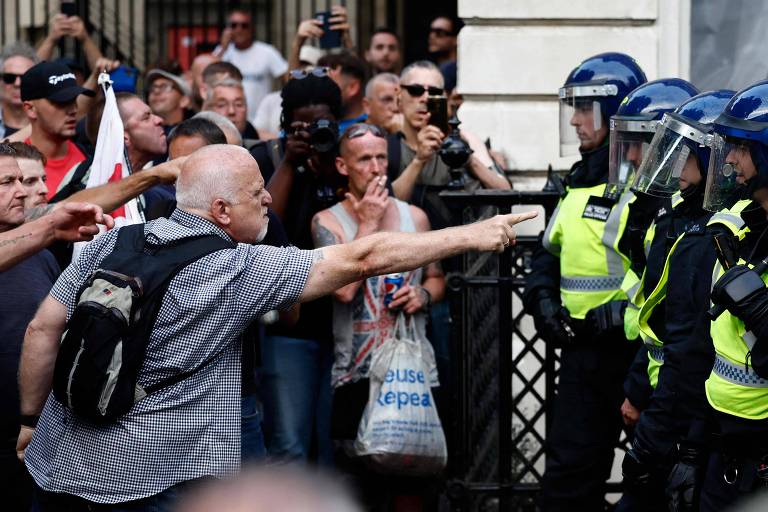 A imagem mostra um grupo de pessoas em uma manifestação, com um homem de camisa xadrez apontando para uma linha de policiais em uniforme de proteção. Os policiais estão em formação, usando capacetes e escudos. Ao fundo, há várias pessoas observando, algumas filmando com celulares. A atmosfera parece tensa.