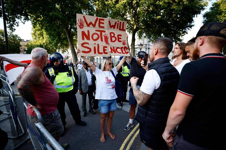 Uma mulher está segurando uma placa que diz 'WE WILL NOT BE SILENCED' em um protesto. Ela está descalça e vestindo uma camiseta branca e shorts. Ao fundo, há um grupo de pessoas, incluindo um policial com uniforme e outros manifestantes. A cena é em uma rua com árvores ao fundo.