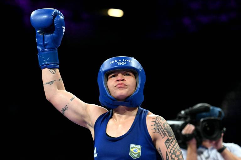 Com capacete, uniforme e luvas azuis, a pugilista Bia Ferreira, com semblante sério, ergue o braço direito após luta nos Jogos Olímpicos de Paris  