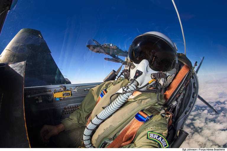 Piloto de caça dentro da cabine de um avião, usando um capacete com visor escuro e uma máscara de oxigênio. O fundo é um céu azul claro com algumas nuvens visíveis. O painel de controle do avião é parcialmente visível à esquerda