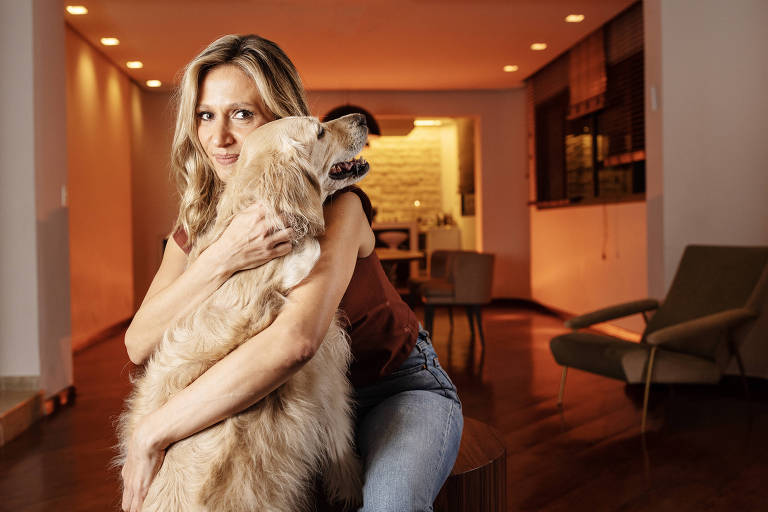 A imagem mostra uma mulher com cabelo longo e liso, vestindo uma blusa vermelha e jeans, abraçando um cachorro de pelagem clara. O ambiente é interno, com iluminação suave e um fundo que sugere uma sala de estar.