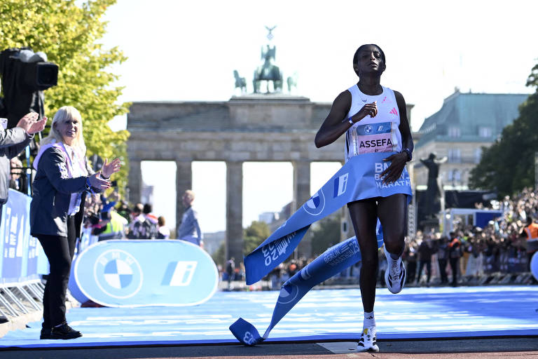 Uma atleta cruzando a linha de chegada da Maratona de Berlim, com um fita azul ao seu redor. Ao fundo, o Portão de Brandemburgo é visível, e há uma multidão aplaudindo. A atleta está vestindo um uniforme de corrida.
