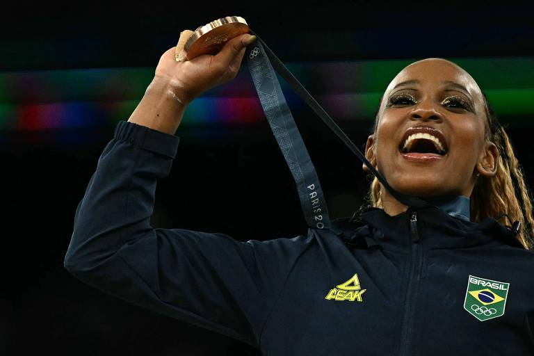 Uma mulher negra é vista dos ombros para cima com uma medalha no pescoço. Ela está sorrindo e está maquiada. O casaco é azul escuro e tem o símbolo da seleção brasileira.