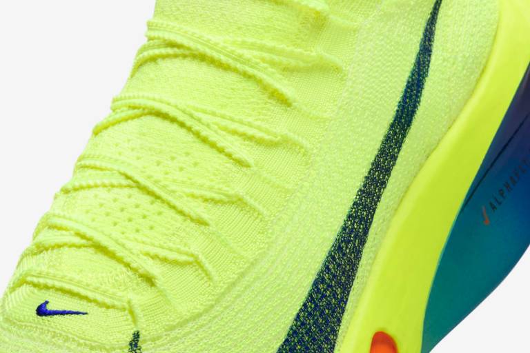 A imagem mostra a parte superior de um tênis da Nike, predominantemente amarelo neon. O cabedal é feito de um material texturizado com detalhes em azul escuro, incluindo o logo da Nike. O solado apresenta cores vibrantes, como azul, laranja e verde, destacando-se na parte traseira do tênis.