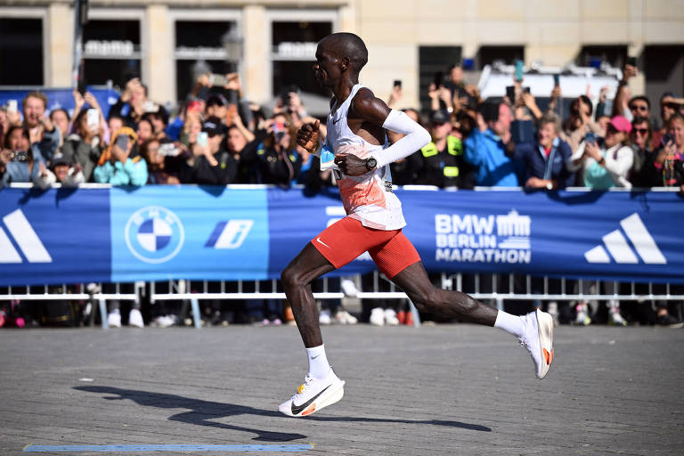 Um corredor está em movimento durante a Maratona de Berlim, com uma multidão de espectadores ao fundo. Ele usa uma camiseta branca com detalhes em vermelho e shorts vermelhos. O corredor parece concentrado e está em uma pose dinâmica, com um pé no ar e o outro no chão. Ao fundo, há uma faixa azul com o logotipo da BMW e da Maratona de Berlim, além de várias pessoas tirando fotos.
