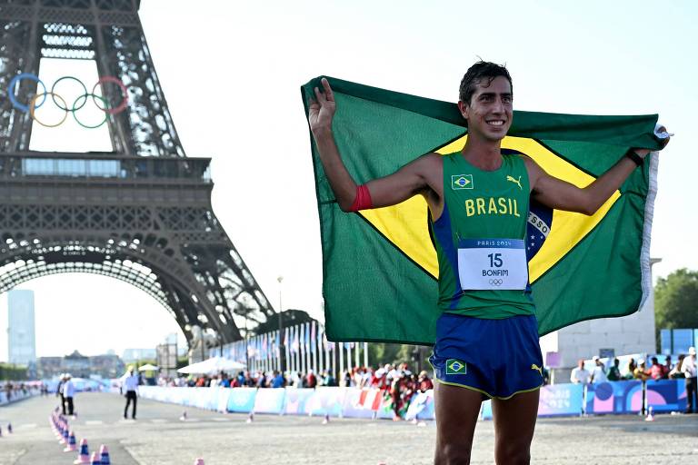 Um atleta brasileiro, vestido com um uniforme verde e amarelo, segura uma bandeira do Brasil enquanto sorri. Ele está posicionado em frente à Torre Eiffel, que é visível ao fundo. O atleta usa um número de identificação, 15, preso ao peito. A cena parece ser de uma competição olímpica.