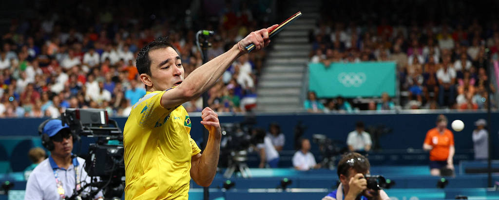 Um atleta está em movimento, saltando e golpeando uma bola de tênis de mesa com uma raquete. Ele veste uma camiseta amarela e shorts pretos. Ao fundo, há uma plateia visível e uma parede com a inscrição 'PARIS 2024'.